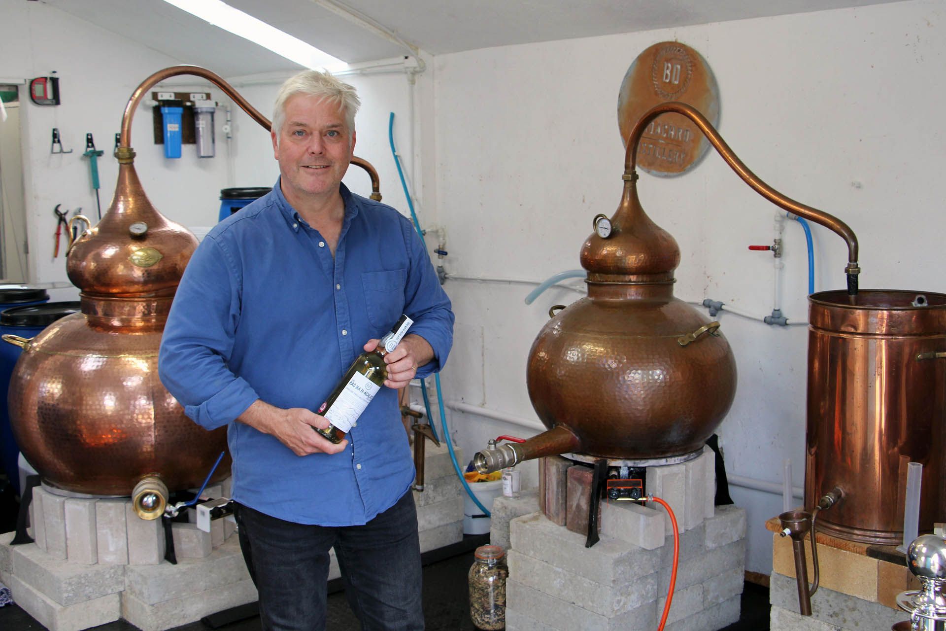 A picture of a man in the Badachro Distillery holding a bottle of whiskey, set next to whisky distillery apparatus in copper
