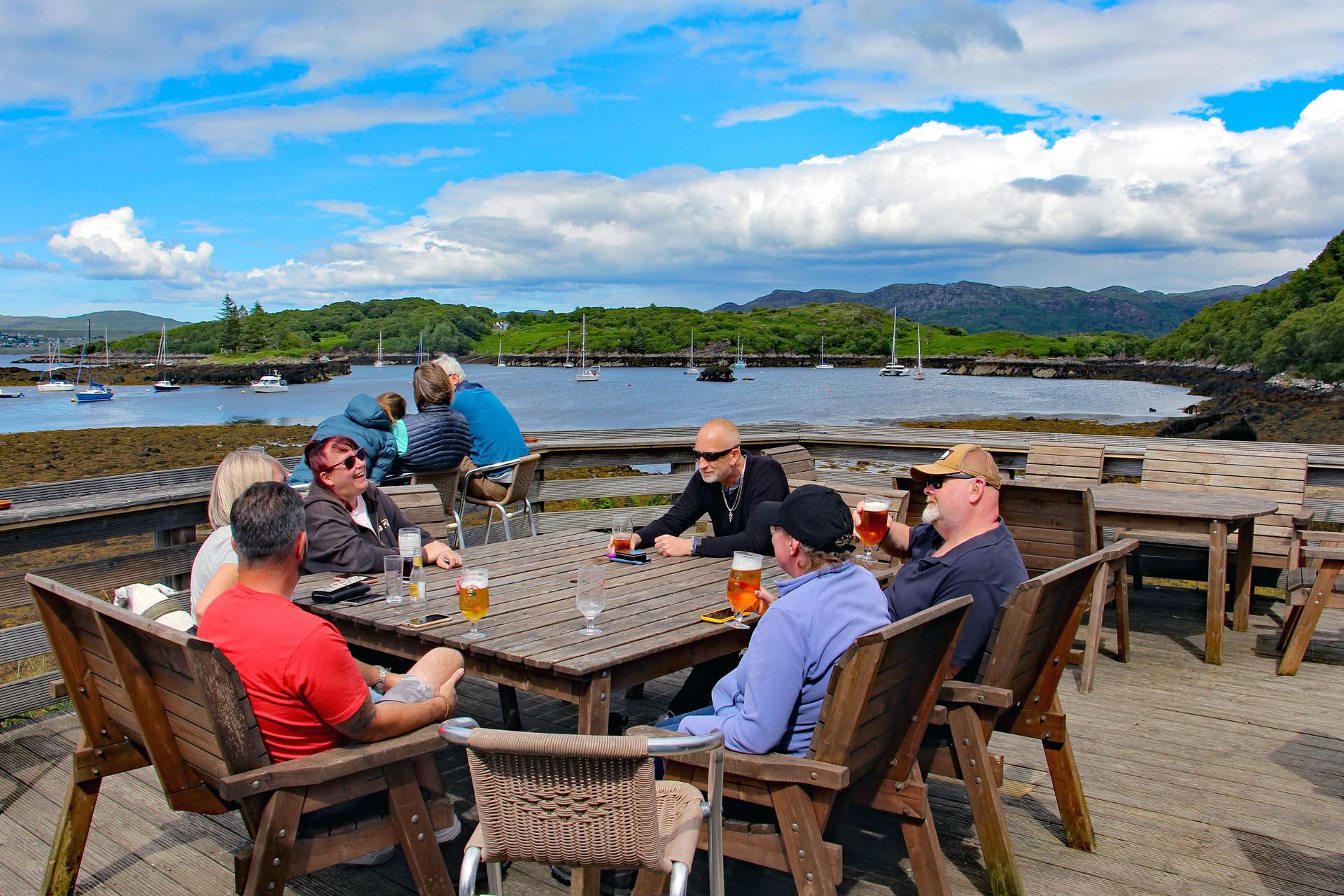 A picture of the outside seating area at the Badachro Inn in Gairloch. The picture includes a group of customers enjoying drinks and good weather. There are boats moored in the background.