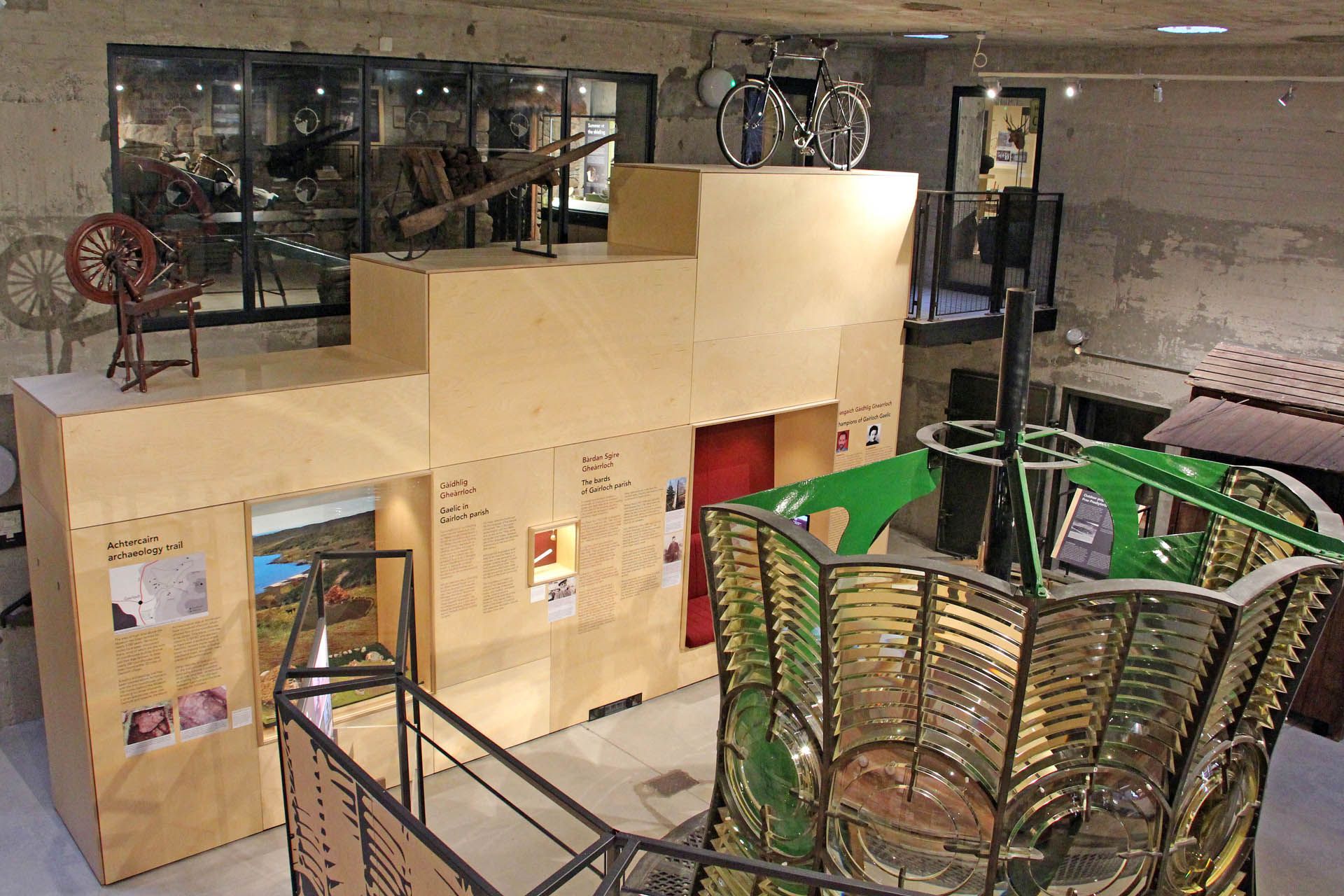A picture of an exhibition within the Gairloch Museum. The picture features exhibits such as a bicycle, spinning wheel and wheel barrow. The image also features the light of a light house